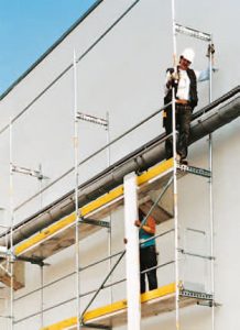 Zwei Arbeiter montieren ein Fassadengerüst an einem Gebäude, eine Person steht auf der oberen Gerüstlage, die andere reicht ein Bauteil von unten an.