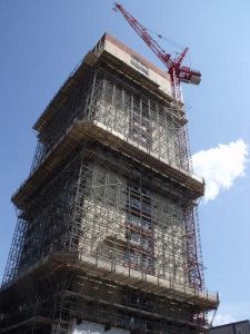 Turmeinrüstung mit mehrlagigem Arbeits- und Schutzgerüst an einem hohen Bauwerk mit rotem Baukran bei strahlend blauem Himmel.