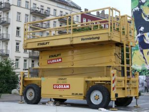 Gelbe mobile Arbeitsbühne vom Typ Scherenbühne auf einem städtischen Platz, vermietet von LOXAM, mit ausgefahrenem Sicherheitsgeländer und abgestellten Stützen vor mehrstöckigen Wohngebäuden.