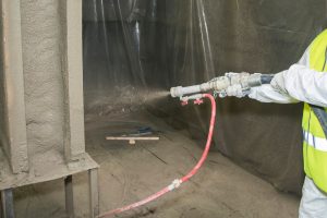 Ein Arbeiter in Schutzkleidung trägt mit einem Sprühgerät eine Betonmischung auf eine Wandoberfläche auf. Das Umfeld ist mit Kunststoffplanen abgedeckt. Ein Werkzeug liegt auf dem Boden. Der Arbeiter trägt eine gelbe Warnweste.