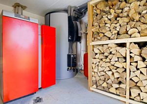Rote Holzheizung und großer Tank stehen in einem Raum. Rechts ist ein Regal mit gestapeltem Brennholz. Die Kombination zeigt eine moderne Nutzung von Holz zur Wärmeerzeugung. Ein kleines Fenster ist zu sehen, ebenso wie Rohrleitungen an der Wand.