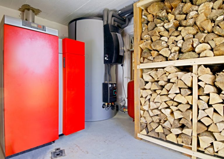 Rote Holzheizung und großer Tank stehen in einem Raum. Rechts ist ein Regal mit gestapeltem Brennholz. Die Kombination zeigt eine moderne Nutzung von Holz zur Wärmeerzeugung. Ein kleines Fenster ist zu sehen, ebenso wie Rohrleitungen an der Wand.