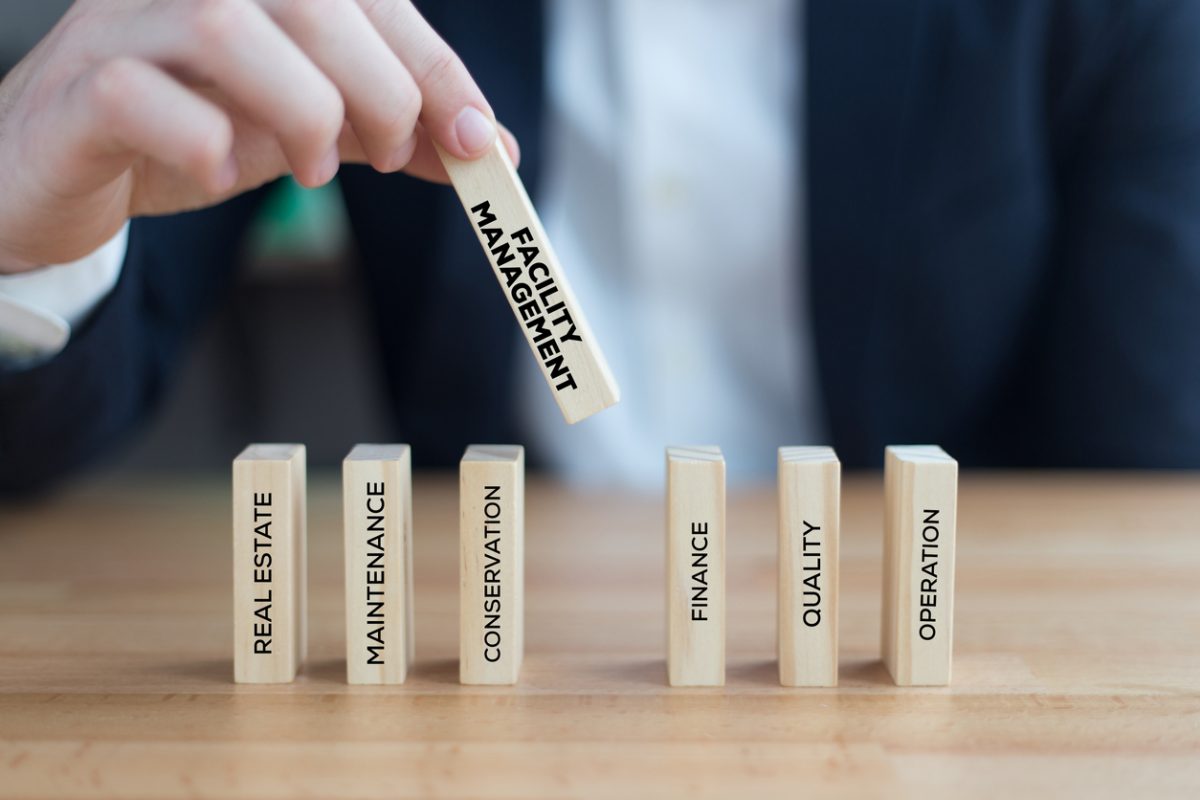 Geschäftsperson hält einen Holzbaustein mit der Aufschrift „Facility Management“ über weiteren Blöcken mit Begriffen wie Real Estate, Maintenance, Conservation, Finance, Quality und Operation – symbolische Darstellung vernetzter Aufgaben im Gebäudemanagement.