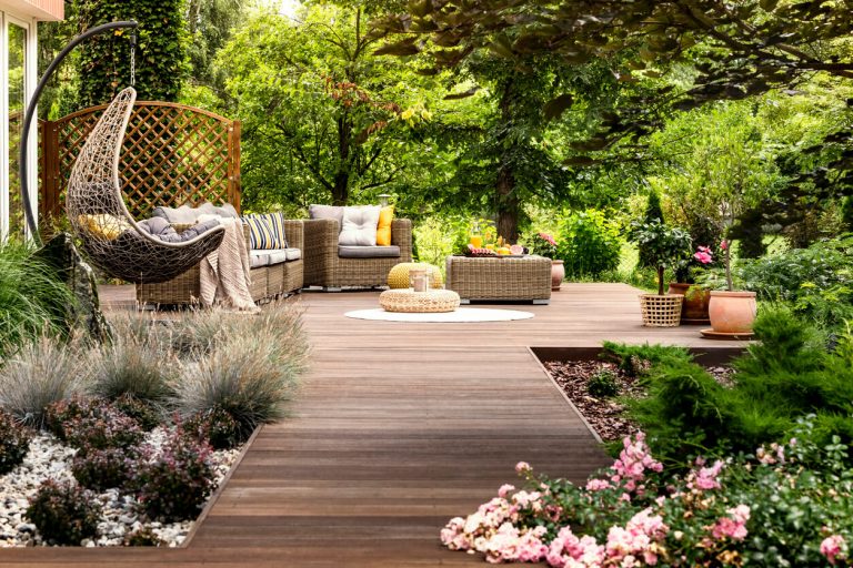 Moderne Holzterrasse mit gemütlichen Rattanmöbeln und üppiger Bepflanzung in einem blühenden Garten.