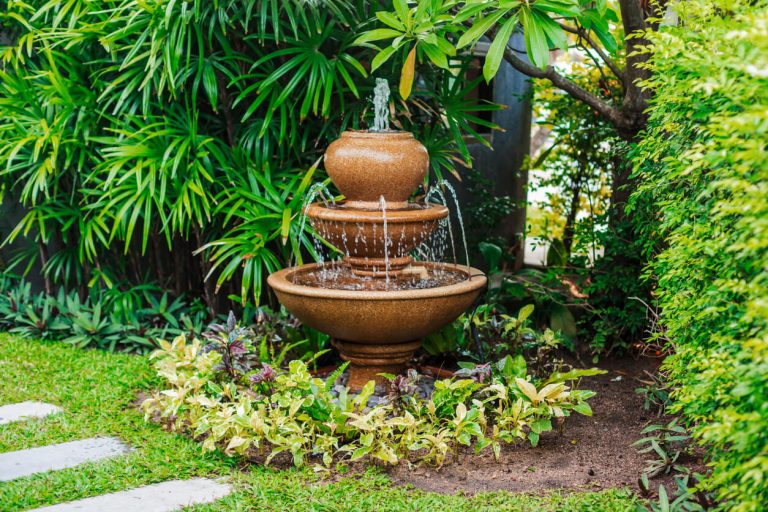 Dreistufiger Zierbrunnen aus Keramik in einem tropisch bepflanzten Garten, umgeben von üppigem Grün und dekorativen Randpflanzen.