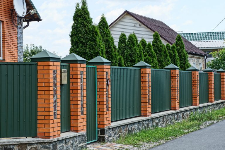 Metall Sichtschutzzaun mit grünen Elementen und roten Backsteinpfeilern vor einem Wohnhaus mit Hecke und Einfahrt