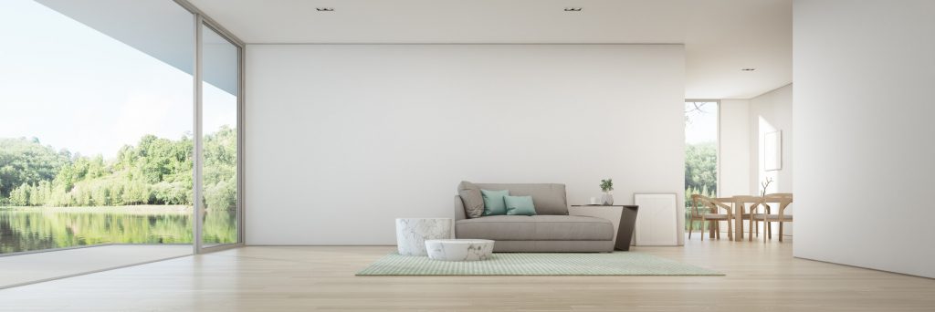 Helles Wohnzimmer mit großer Fensterfront und Blick auf Wald und See. Im Raum steht ein graues Sofa mit grünen Kissen auf einem hellgrünen Teppich, daneben zwei kleine Tische. Im Hintergrund ist ein Esstisch mit Stühlen zu sehen.