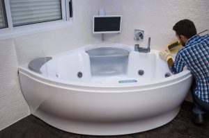 Eine Person in kariertem Hemd und mit Handschuhen führt Wartungsarbeiten an einer großen, weißen, modernen Badewanne mit eingebautem Bildschirm durch. Die Person trägt Dichtmittel entlang der Kante auf.