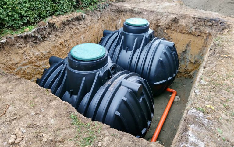 Zwei große schwarze Kunststofftanks sind in einem Erdaushub installiert. Sie sind durch ein orangefarbenes Rohr verbunden und mit grünen Abdeckungen versehen. Die Grube ist von Erdreich umgeben, und im Hintergrund sind Büsche zu sehen.