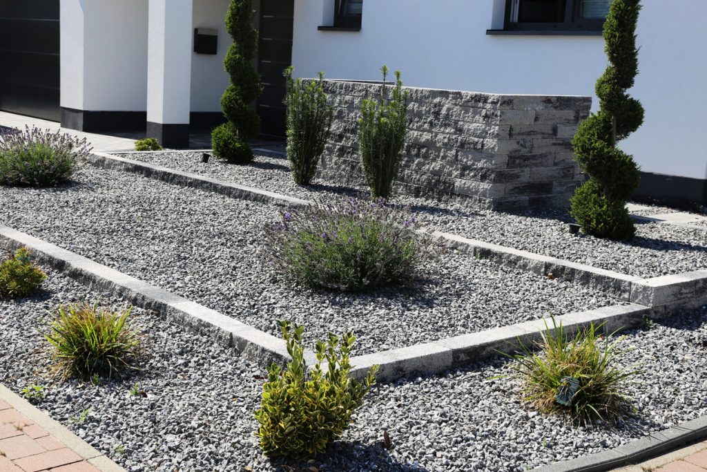 Moderner Vorgarten mit Schotterfläche, vereinzelten Zierpflanzen und dekorativen Gehölzen vor einem weißen Einfamilienhaus.