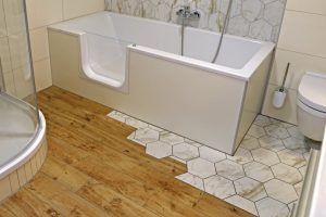 Badezimmer mit einer Badewanne, die einen seitlichen Einstieg hat. Der Boden ist teils aus Holzoptikfliesen und teils aus sechseckigen Marmorfliesen. Eine Dusche und eine Toilette sind ebenfalls sichtbar.