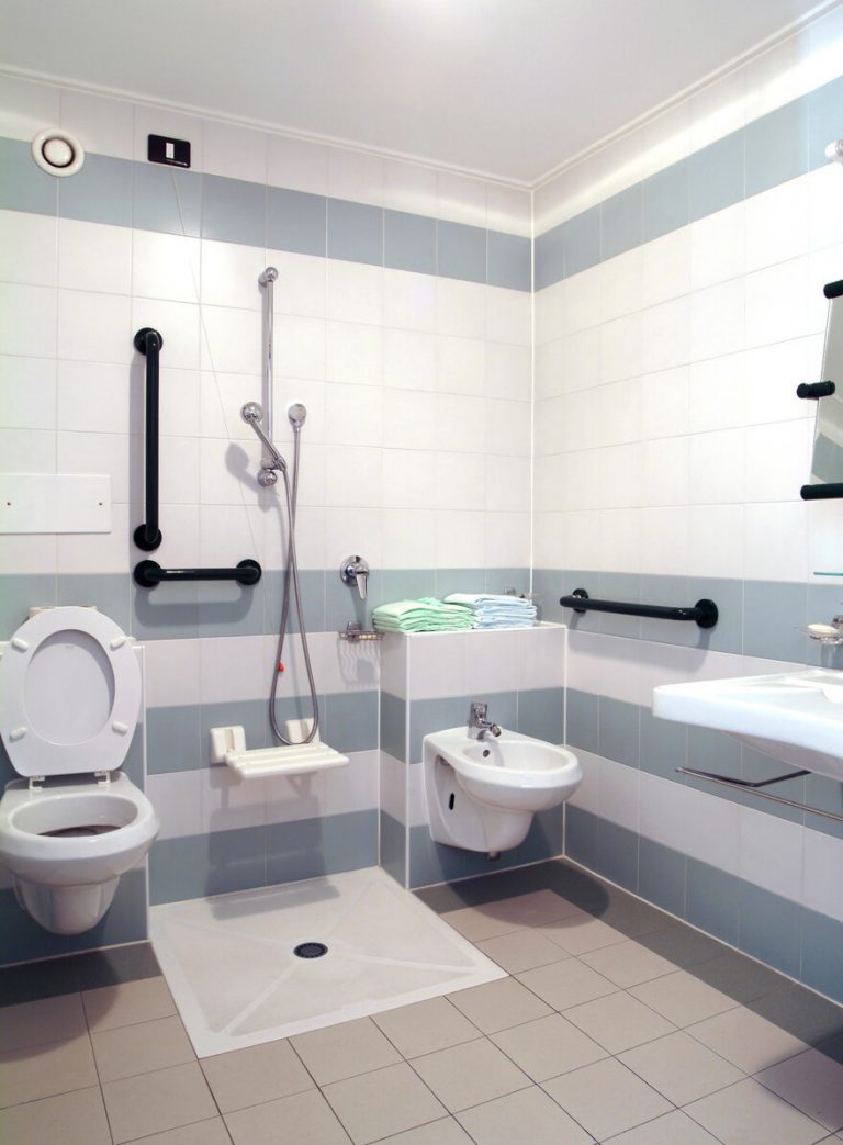 Ein barrierefreies Badezimmer mit einer bodengleichen Dusche, vielzähligen Haltegriffen, einem WC, einem Waschbecken und einem Bidet. Handtücher sind auf einem Regal gestapelt. Die Fliesen sind in Weiß und Grau gehalten.
