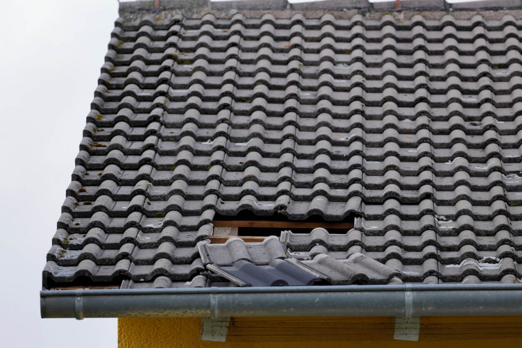 Dach mit grauen Ziegeln, eine Reihe von Ziegeln fehlt und das darunterliegende Holz ist sichtbar. Eine Dachrinne verläuft am unteren Rand. Der Himmel ist bewölkt.