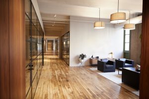 Modernes Büro mit Parkettboden, der eine warme, natürliche Atmosphäre schafft. Der Flur ist von Glaswänden gesäumt und der Wartebereich mit schwarzen Sesseln bietet eine stilvolle, minimalistische Einrichtung.