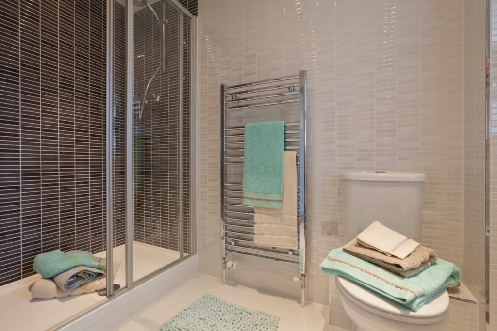 Modernes Badezimmer mit Glastür-Dusche, chromfarbenem Handtuchhalter, auf dem türkisfarbene Handtücher hängen. Ein Stapel Handtücher liegt auf der Toilette, der Boden ist mit einer türkisfarbenen Badematte bedeckt. Wandfliesen in Braun und Weiß.