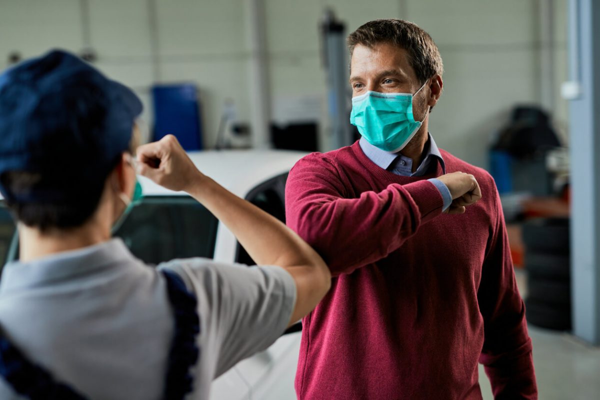 Zwei Männer mit Mundschutz begrüßen sich kontaktlos mit einem Ellenbogengruß in einer Werkstattumgebung zur Einhaltung von Hygieneregeln.