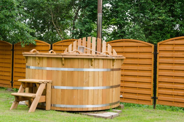 Holzbefeuerter Hot Tub aus Holz im Garten, mit Holztreppe und Sichtschutzwand im Hintergrund.