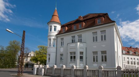 Weiße Stadtvilla mit rotem Mansarddach, das zwei unterschiedlich steile Dachflächen pro Seite aufweist. In das steilere untere Dachsegment sind mehrere Gauben mit kleinen Fenstern eingebaut. Der Gebäudestil erinnert an die Gründerzeit, mit dekorativen Fassadenelementen und einem runden Eckturm mit spitzem Kegeldach.