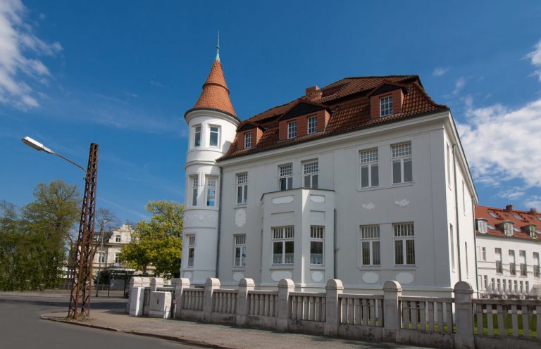 Weiße Stadtvilla mit rotem Mansarddach, das zwei unterschiedlich steile Dachflächen pro Seite aufweist. In das steilere untere Dachsegment sind mehrere Gauben mit kleinen Fenstern eingebaut. Der Gebäudestil erinnert an die Gründerzeit, mit dekorativen Fassadenelementen und einem runden Eckturm mit spitzem Kegeldach.