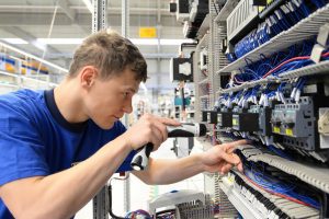Ein junger Informationselektroniker arbeitet konzentriert an einem Schaltschrank. Er verwendet einen Akkuschrauber, um elektrische Komponenten zu montieren oder zu justieren. Die zahlreichen blauen Kabel deuten auf eine komplexe Elektroinstallation hin.