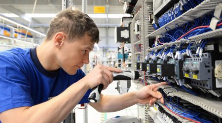 Ein junger Informationselektroniker arbeitet konzentriert an einem Schaltschrank. Er verwendet einen Akkuschrauber, um elektrische Komponenten zu montieren oder zu justieren. Die zahlreichen blauen Kabel deuten auf eine komplexe Elektroinstallation hin.