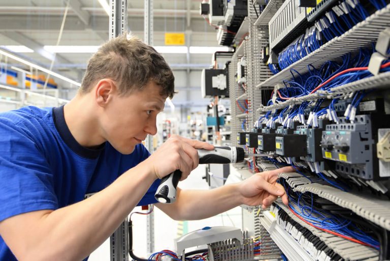 Ein junger Informationselektroniker arbeitet konzentriert an einem Schaltschrank. Er verwendet einen Akkuschrauber, um elektrische Komponenten zu montieren oder zu justieren. Die zahlreichen blauen Kabel deuten auf eine komplexe Elektroinstallation hin.