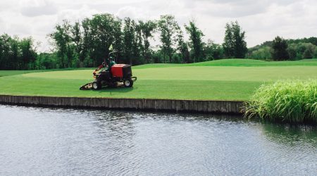 Greenkeeper mäht mit Rasenmäher das Grün eines Golfplatz am Wasser