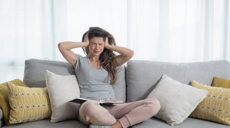 Junge Frau auf einem grauen Sofa presst sich beide Hände auf die Ohren wegen Ohrgeräuschen