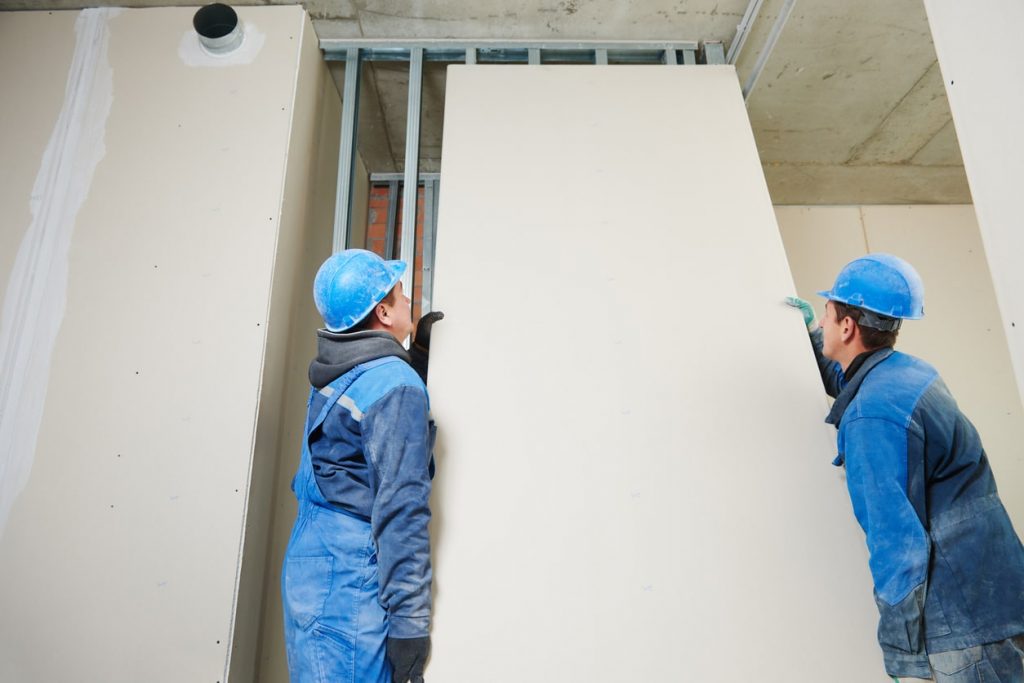 Zwei Handwerker mit blauen Helmen setzen die Gipskartonplatten auf die Profile einer Trockenbauwand