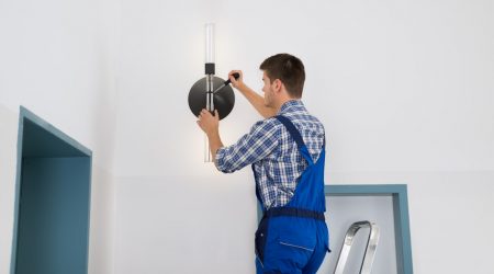 Hausmeister mit Blaumann montiert eine Deckenlampe auf einer Leiter