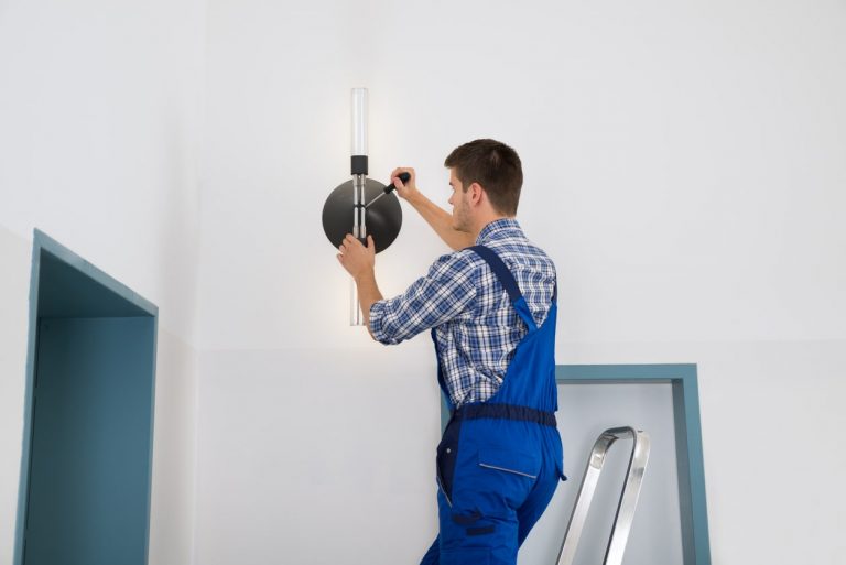 Hausmeister mit Blaumann montiert eine Deckenlampe auf einer Leiter