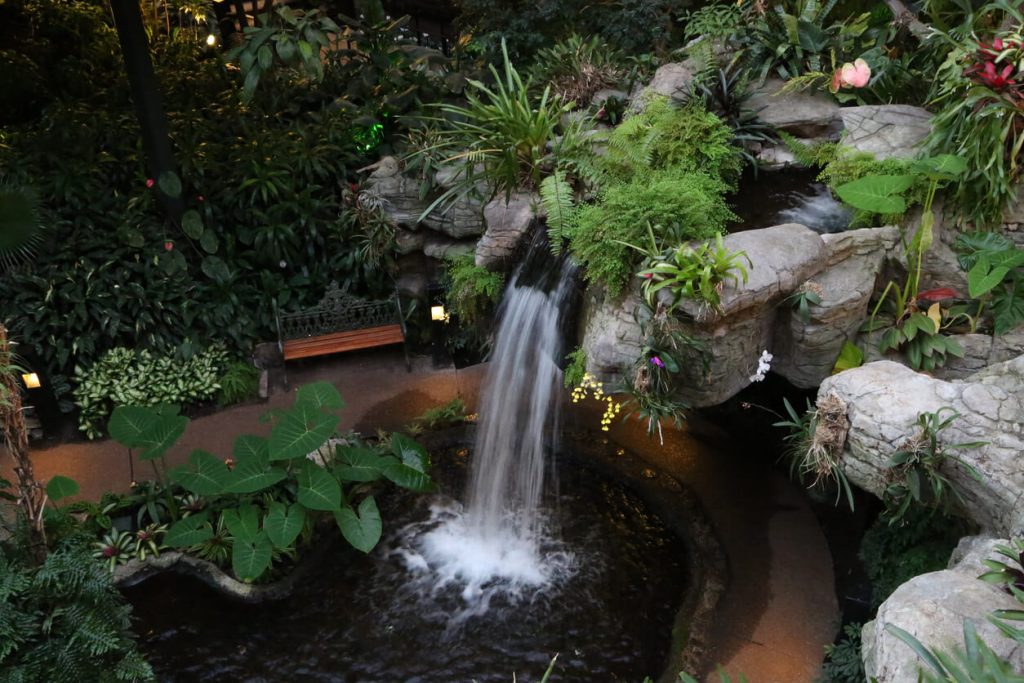 Großer künstlicher Wasserfall in tropischem Gartenambiente mit üppiger Bepflanzung, Natursteinen, Teich und Sitzbank im Hintergrund.
