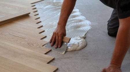 Bodenleger bringt Kleber für Holzboden mit einer Spachtel auf dem Boden auf