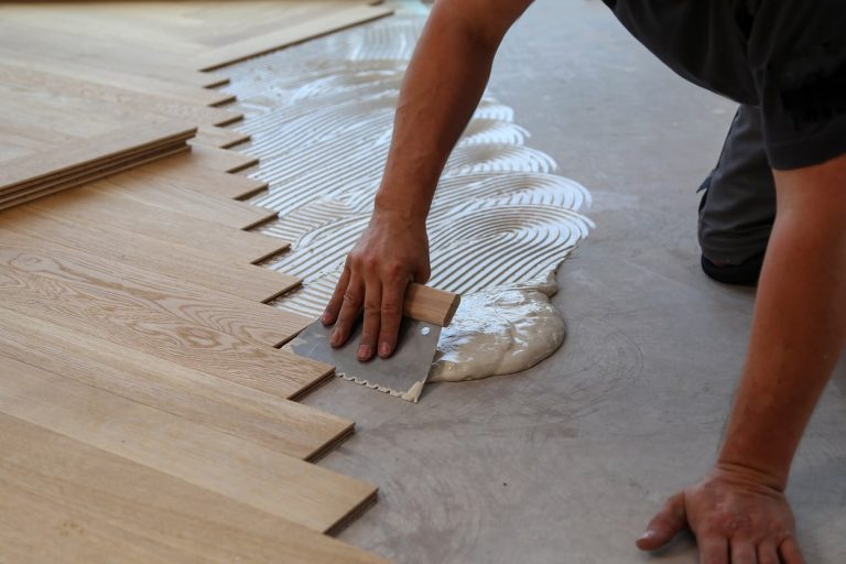 Bodenleger bringt Kleber für Holzboden mit einer Spachtel auf dem Boden auf