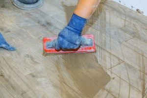 Fliesenleger mit blauen Gummihandschuhen bringt Fugenmasse auf den Boden auf