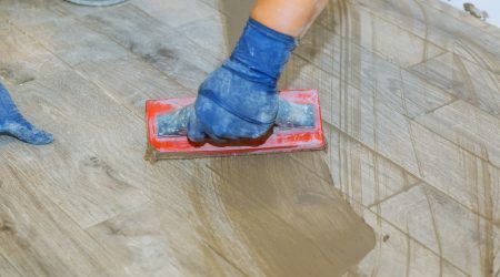 Fliesenleger mit blauen Gummihandschuhen bringt Fugenmasse auf den Boden auf
