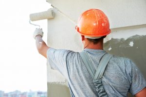 Maler mit Farbroller in der linken Hand und orange farbigem Bauhelm streicht die Fassade eine Gebäudes