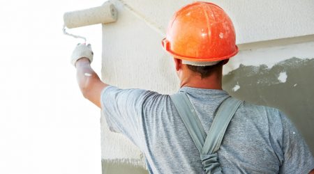 Maler mit Farbroller in der linken Hand und orange farbigem Bauhelm streicht die Fassade eine Gebäudes