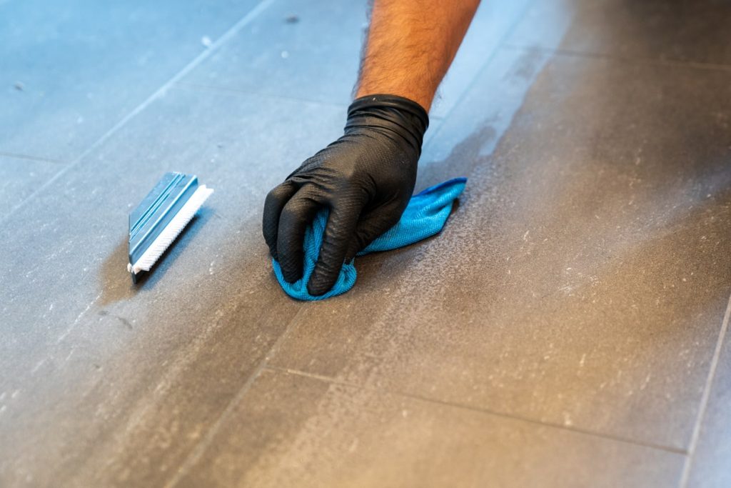 Fliesenleger mit schwarzen Gummihandschuhen reinigt Fugen und Fliesen mit Reiniger nach dem Verfugen
