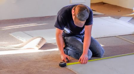 Bodenleger mit Jeans und Mütze verlegt einen Teppich mit Maßband und Teppichmesser in einem Raum