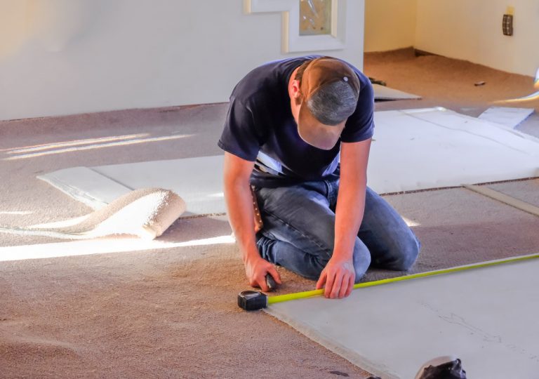 Bodenleger mit Jeans und Mütze verlegt einen Teppich mit Maßband und Teppichmesser in einem Raum