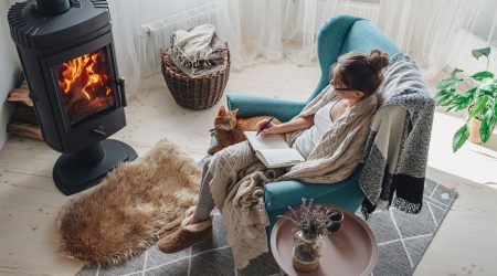 Junge Frau sitzt mit Decke und Notizbuch auf einem Sessel vor einem Kaminofen