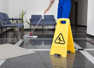 Hausmeister reinigt mit einem Mob ein öffentliches Gebäude mit einem gelbem Achtung Rutschgefahr Schild im Vordergrund