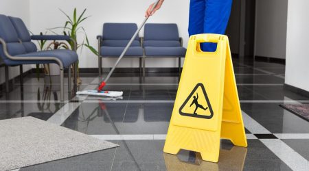 Hausmeister reinigt mit einem Mob ein öffentliches Gebäude mit einem gelbem Achtung Rutschgefahr Schild im Vordergrund