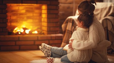Mutter sitzt mit Tochter umarmend vor einem brennenden Kamin auf einem Fell