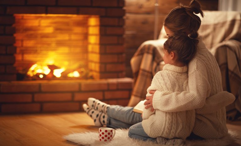 Mutter sitzt mit Tochter umarmend vor einem brennenden Kamin auf einem Fell