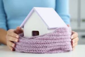Frauenhände halten ein in einen lila farbigen, gestrickten Schal eingewickeltes kleines Holzhaus als Symbol zur Wärmedämmung