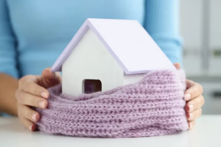 Frauenhände halten ein in einen lila farbigen, gestrickten Schal eingewickeltes kleines Holzhaus als Symbol zur Wärmedämmung