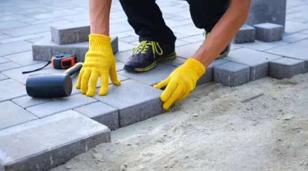 Terrassenbauer mit gelben Handschuhen verlegt graue Betonpflasterplatten im Haushof auf Sandfundamentsockel