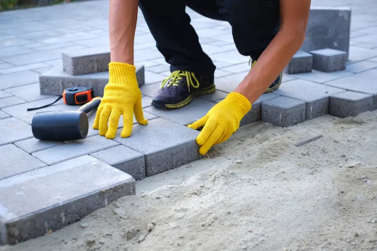 Terrassenbauer mit gelben Handschuhen verlegt graue Betonpflasterplatten im Haushof auf Sandfundamentsockel